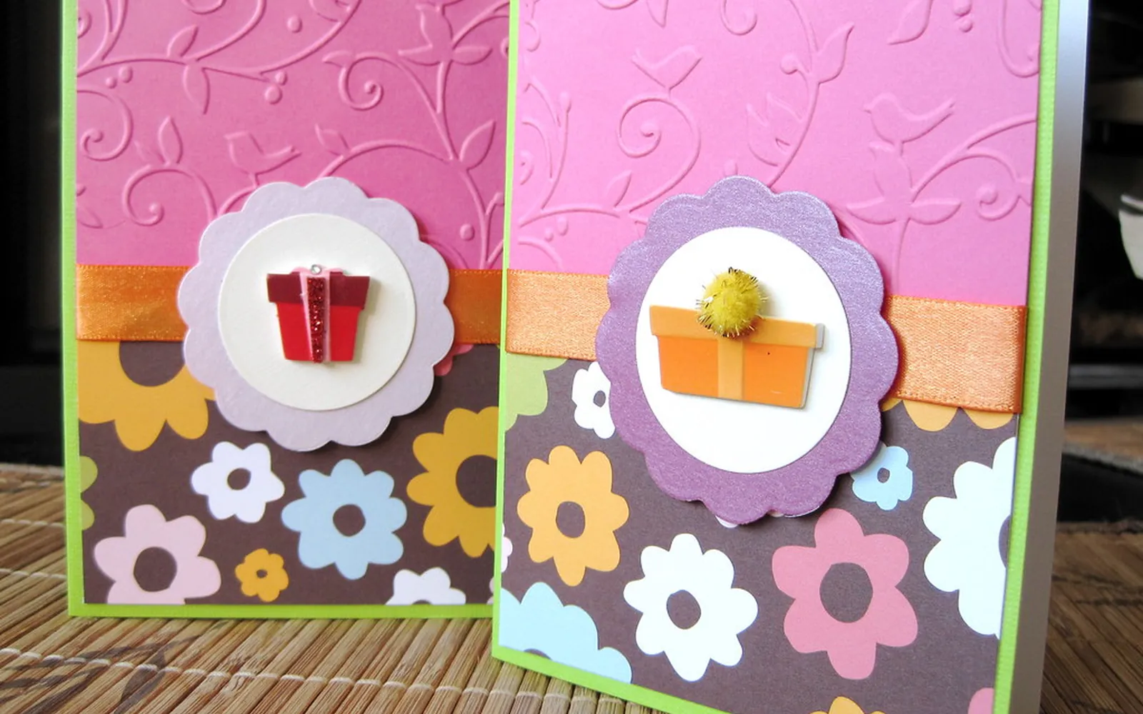Two handmade greeting cards with pink embossed tops, ribbon and flower-pattern paper standing on a table