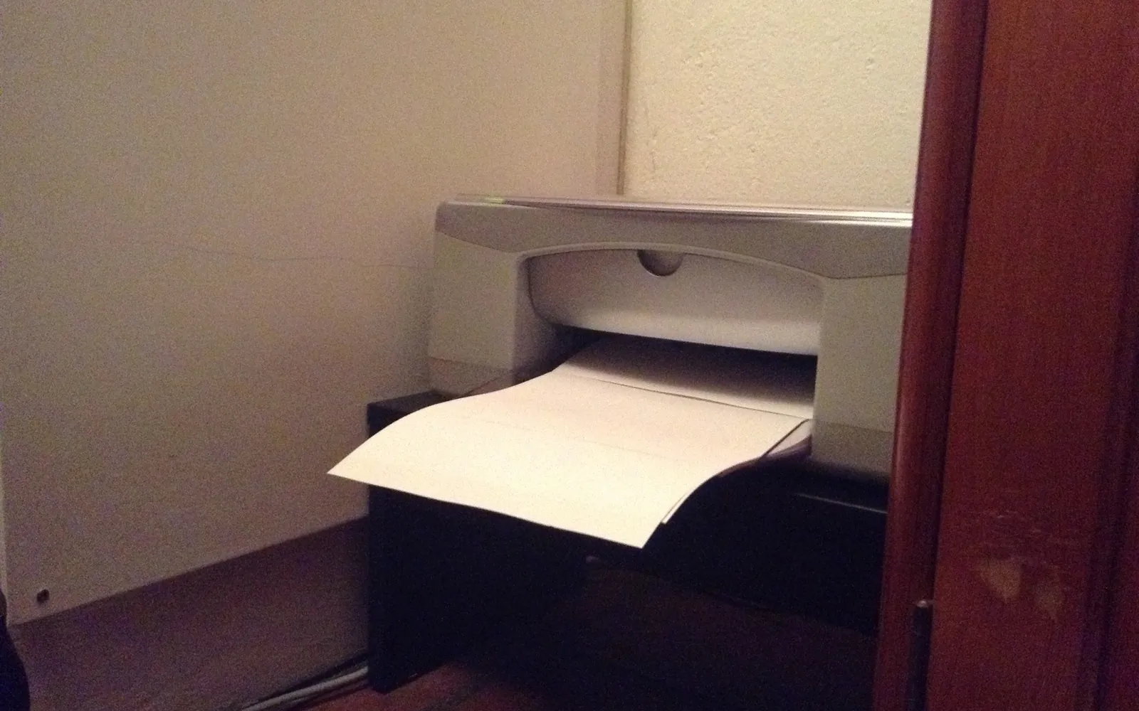 A small all-in-one inkjet printer on a shelf in the corner of a room, with paper in the output tray
