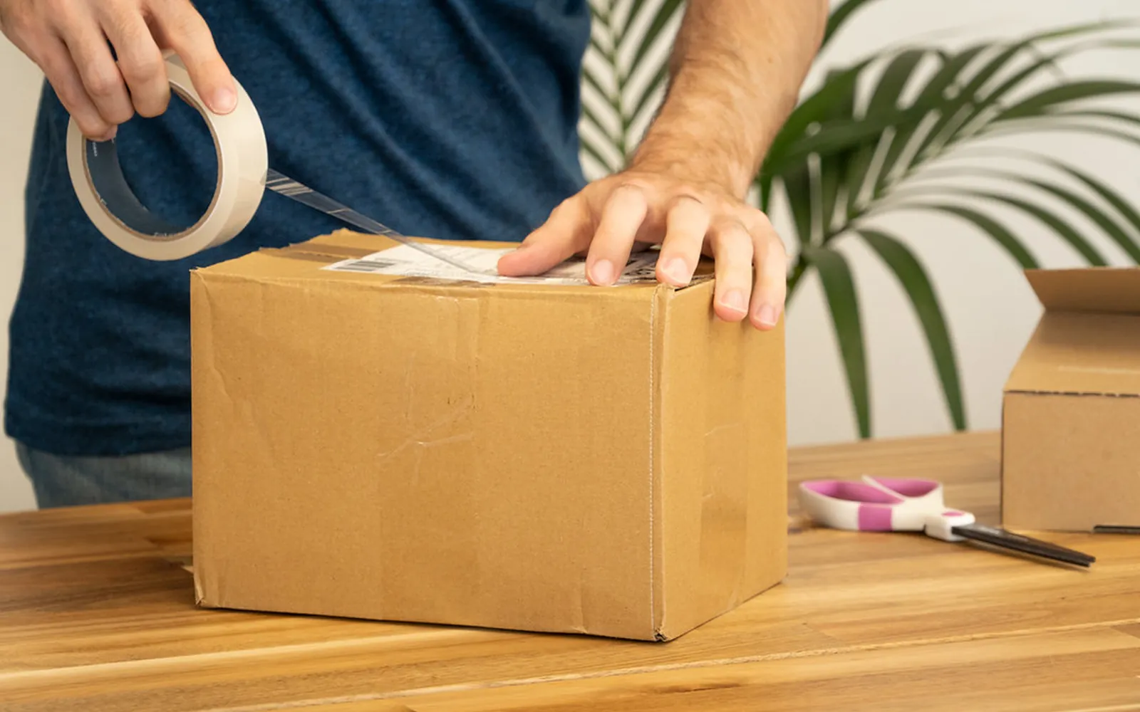 A person sealing a cardboard parcel with packing tape on a wooden table, ready for a printed shipping label