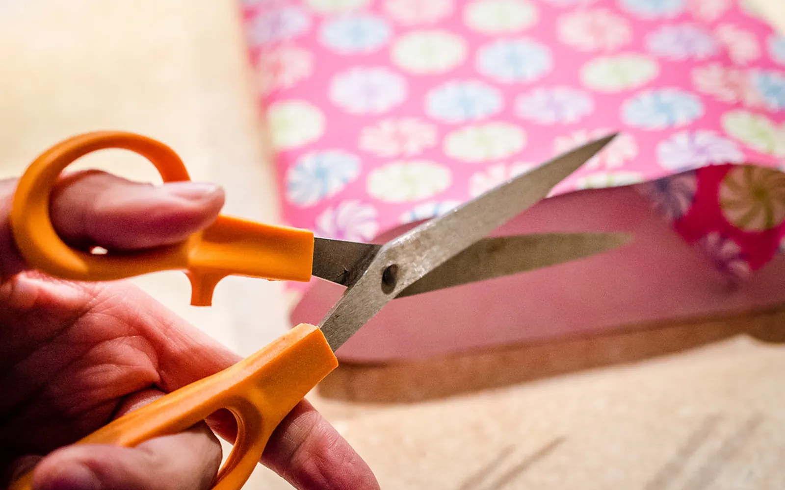 Hands cutting a sheet of patterned paper with orange-handled scissors, the same way home-printed stickers are trimmed