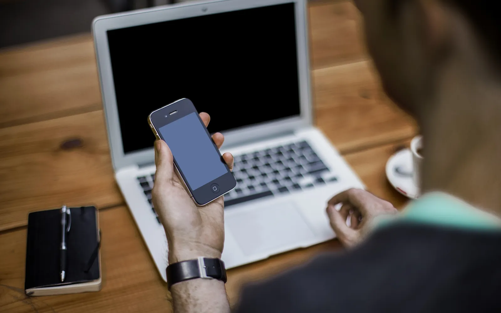 A person holding a smartphone above a laptop on a wooden desk