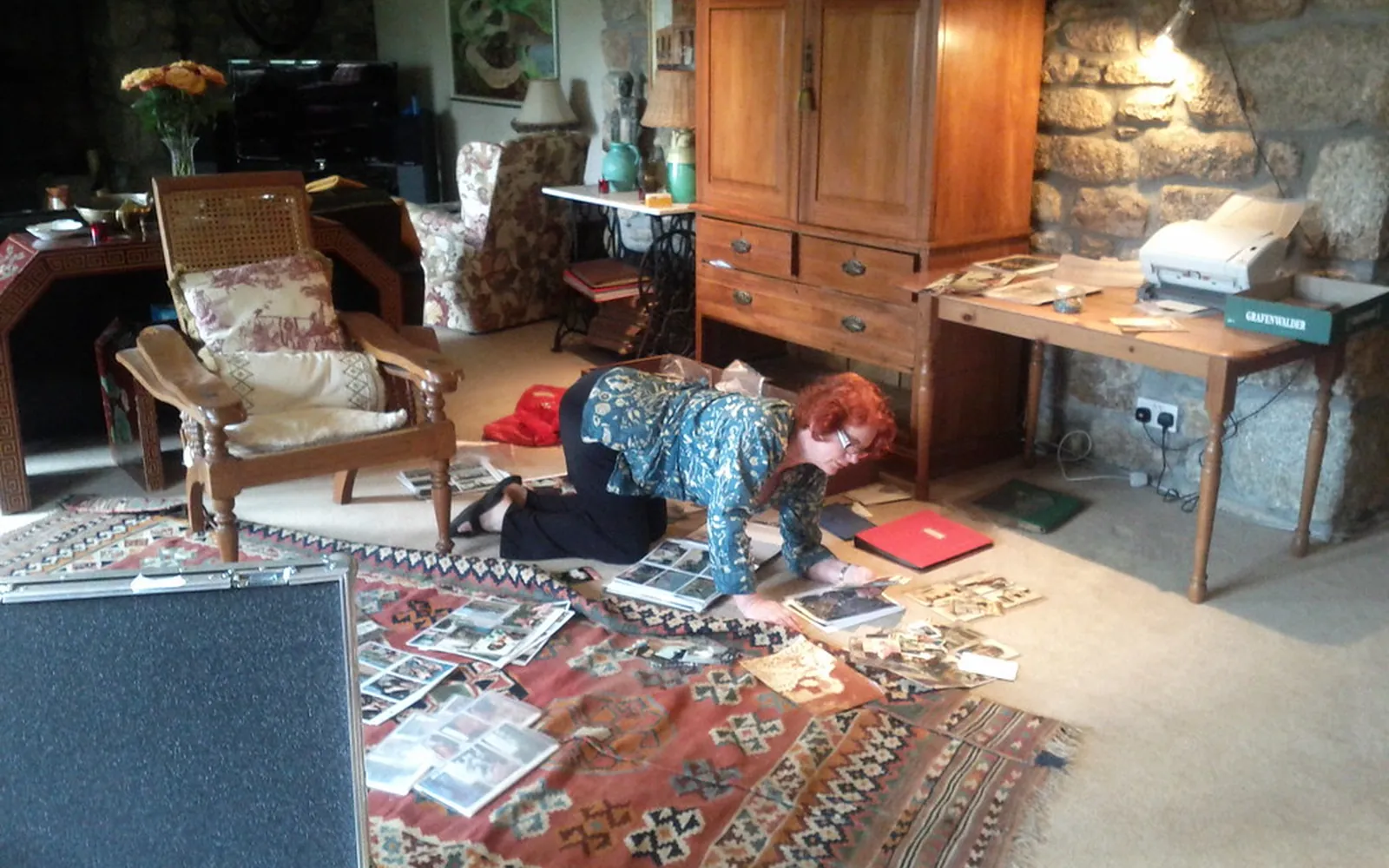 A person sorting piles of old family photographs and albums on a living room rug, with a printer on a side table